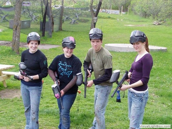 Four people holding paintball guns stand in a grassy park with trees and benches.