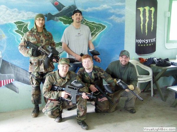 Five camouflaged people posing with airsoft guns in an indoor room with a tropical mural and Monster poster