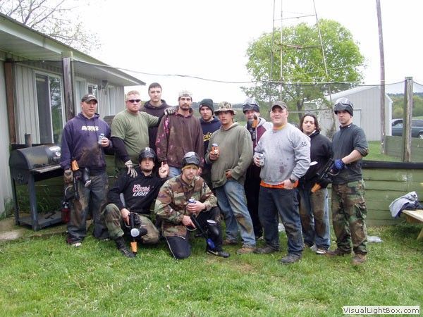 Group of people posing outdoors in camouflage and casual clothes beside a house, with one kneeling in front.