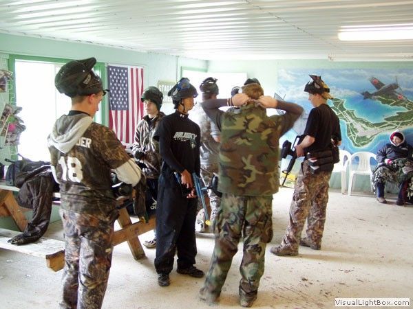 Uniformed personnel gathered indoors in a room with a U.S. flag and wall mural, adjusting gear and talking.