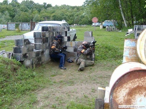 Two people in tactical gear crouch behind stacked concrete blocks near a van in a grassy field.