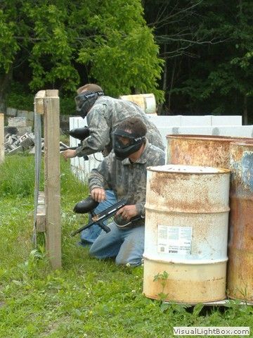 Two people in camouflage crouch behind barrels and a post, aiming paintball guns outdoors.