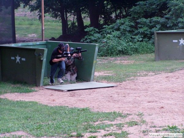 Two people crouch in a green paintball bunker aiming markers on a grassy outdoor field.