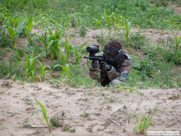 Person in camouflage crouches with a paintball gun in a grassy field.