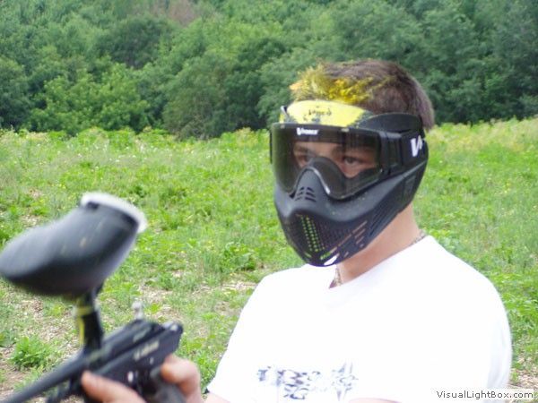 Person wearing a paintball mask and white shirt holding a paintball gun outdoors on a grassy field