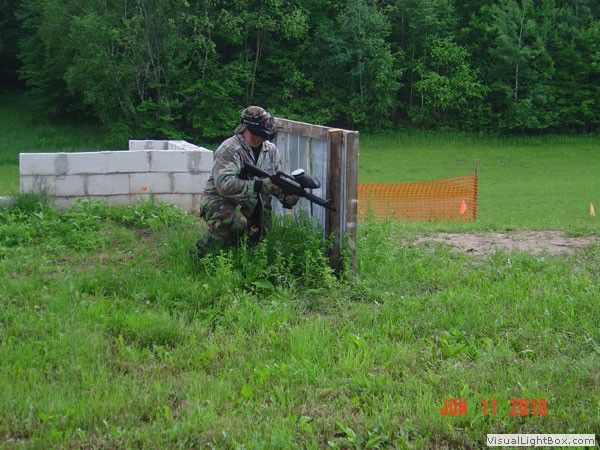 Soldier crouched behind a concrete barrier aiming a rifle on a grassy field with orange safety fencing