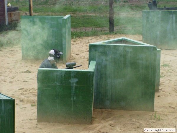 Person in helmet crouches behind green paintball barriers in a dusty outdoor arena.