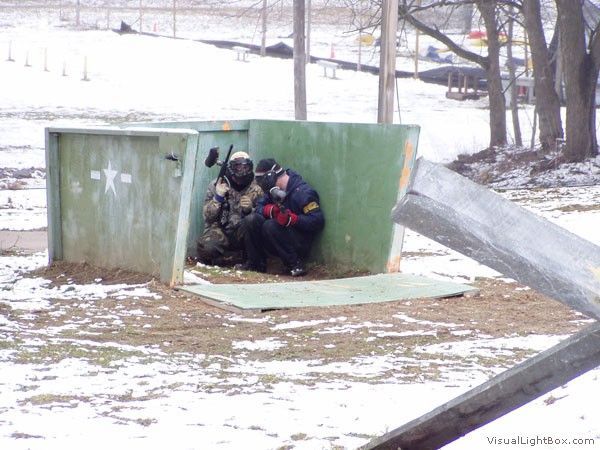Two people crouch in a green bunker entrance in a snowy outdoor training area.