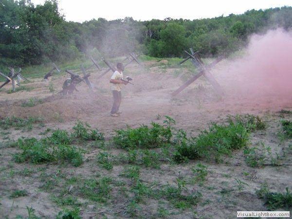 Person in a field amid pink smoke, with dust and greenery in the background