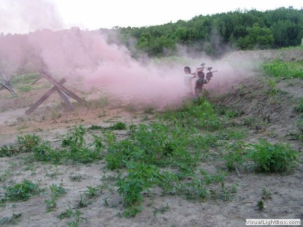 Person spraying pink smoke in a grassy field near a wooded hillside