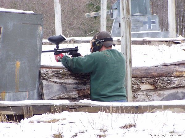 Person aiming a paintball gun behind a wooden barricade in a snowy outdoor field