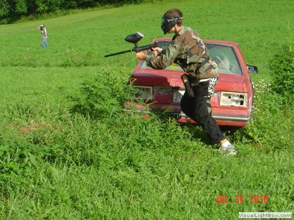 Person in camouflage aiming a paintball marker beside a red car in a grassy field.