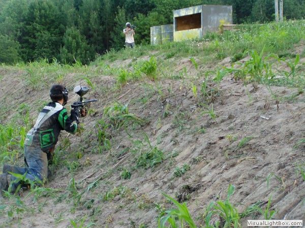 Person crouches on a grassy hillside aiming a paintball gun toward a target near a small shed.