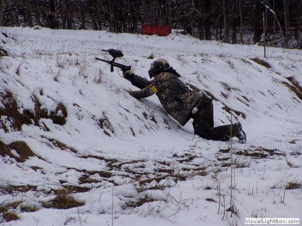 Camouflaged person kneels in snow aiming a paintball gun at a snowy hillside
