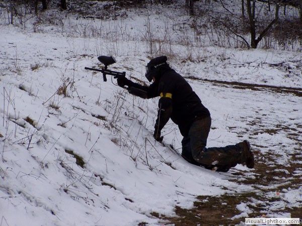 Person kneeling in snow aiming a paintball gun on a snowy hillside