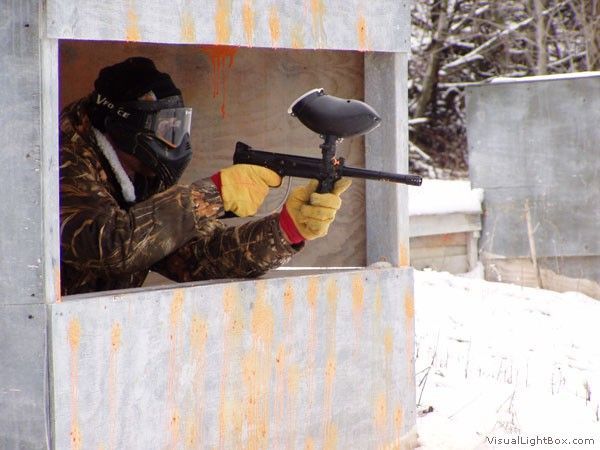 Person in paintball mask aiming a marker from a wooden bunker in a snowy field.