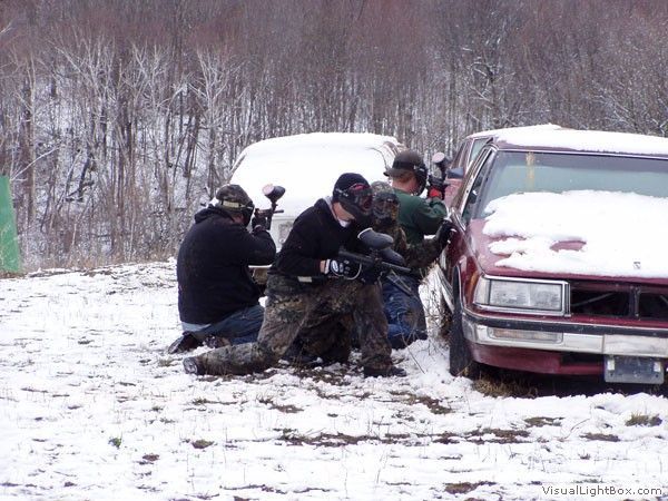 Four people push a red SUV stuck in snow on a muddy roadside near a wooded area.