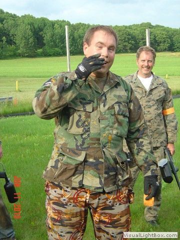 Two men in camouflage outdoors; one waves at the camera on a grassy field.