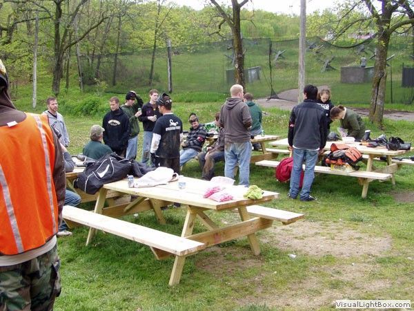 People gathered around picnic tables in a park, with bags and food spread out on benches.