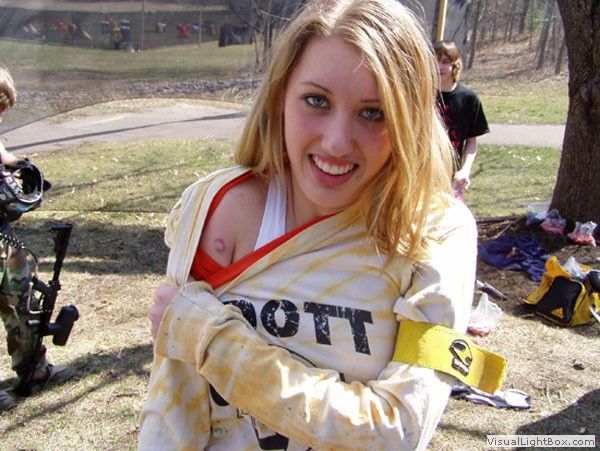 Smiling woman in a white top with orange straps outdoors, holding a camera and wearing a yellow armband.