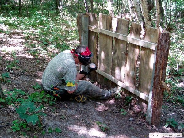 Person kneeling by a wooden fence, using a saw in a wooded yard.