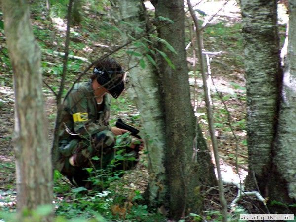 Camouflaged person crouches in a forest among trees, wearing a dark helmet and holding equipment.