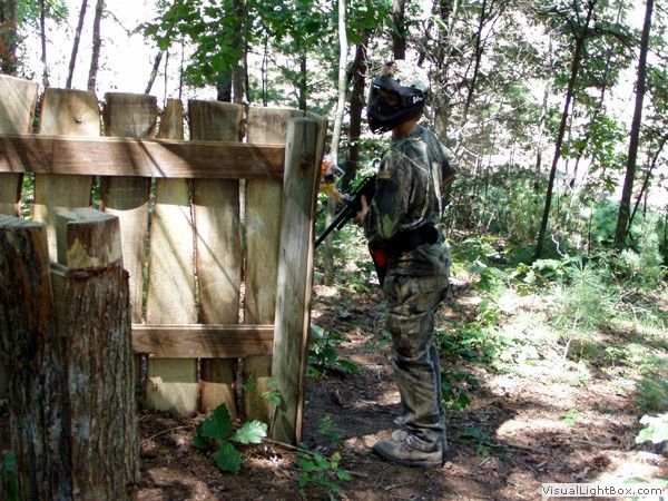 Person in camouflage stands beside a wooden barrier in a wooded outdoor setting.
