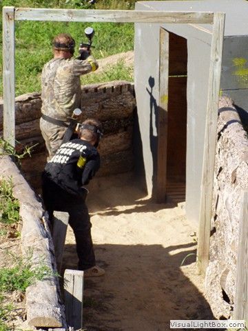 Two people in tactical gear aim paintball guns at a wooden bunker doorway outdoors.