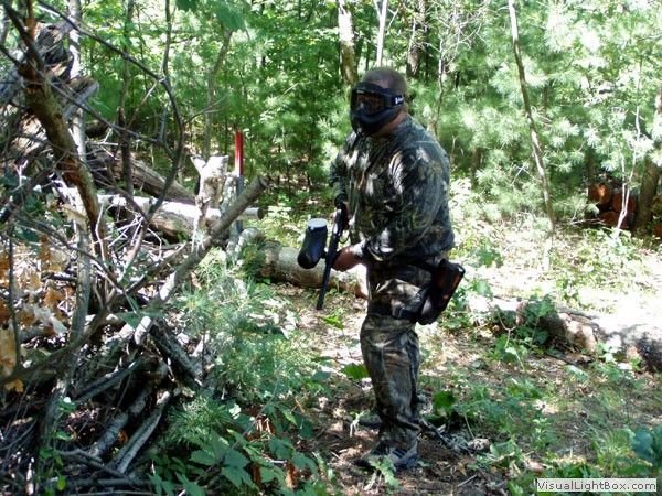 Camouflaged person holding a paintball marker in a wooded outdoor area