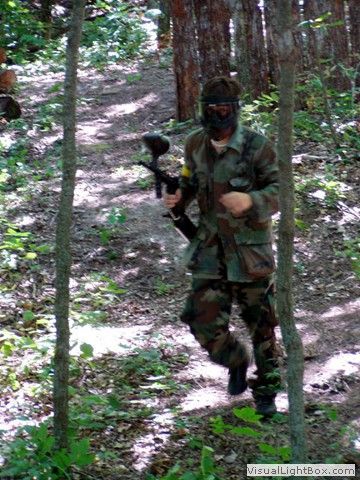 Camouflaged person holding a paintball gun walking through a wooded trail