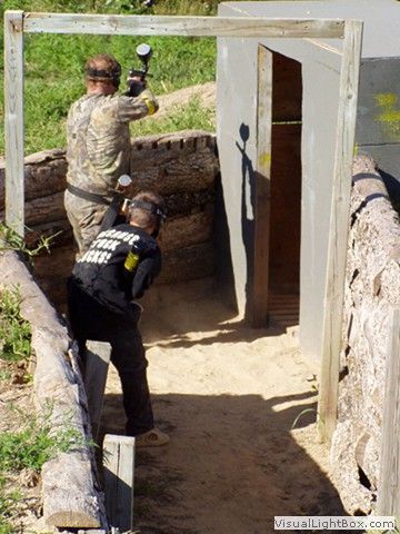 Two people in paintball gear aim markers at a target behind a concrete barrier.