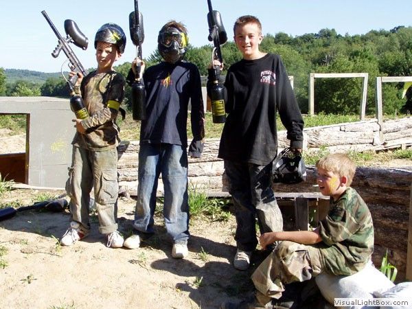 Four people in camouflage gear pose outdoors holding paintball guns near a wooden structure.