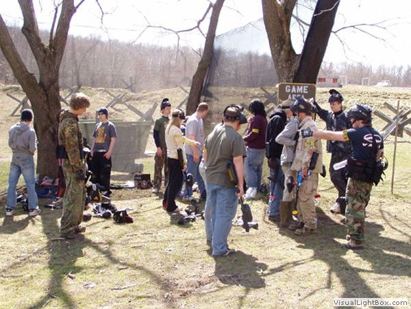 People gathered outdoors in a wooded area, some holding gear and talking near a sign marked “GAME.”