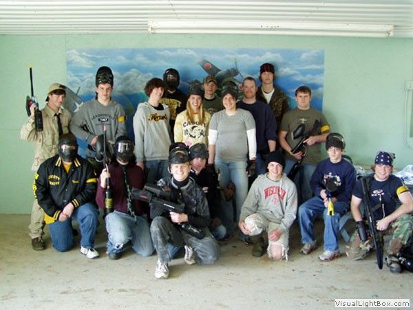 Group of people in paintball gear posing indoors, with a mural backdrop and fluorescent lights.