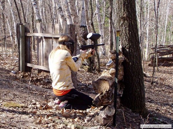 Person kneeling in a wooded area, using a power tool on a log beside a fence and trees