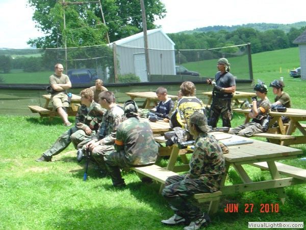 Camouflage-clad group seated at picnic tables outdoors near a white shed on a sunny day