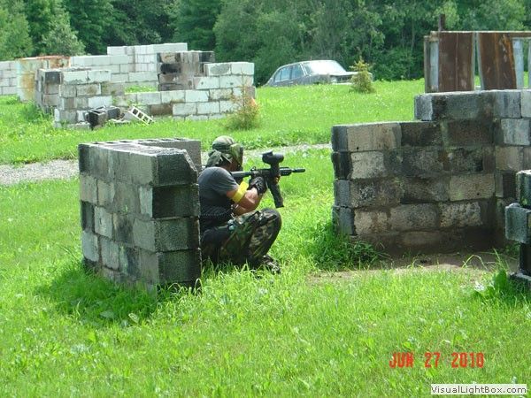 Person in tactical gear crouches behind a concrete barrier aiming a paintball marker on a grassy field.