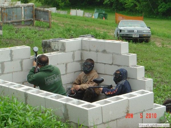 Three people taking cover in a cinderblock bunker, one aiming a marker in a grassy field.