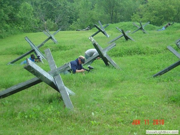 Person crawling under metal X-shaped obstacles on a grassy outdoor obstacle course.