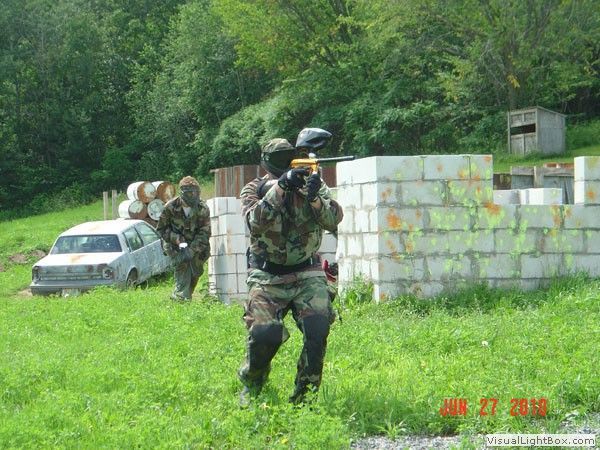 Camouflaged people practice near a white car and concrete barricades in a grassy wooded area.