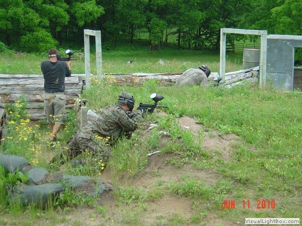 Two people in camouflage aiming paintball guns in an outdoor field with bunkers and grass.