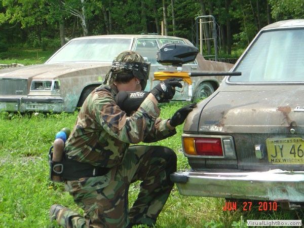 Camouflaged person crouches and aims a paintball marker beside a parked sedan in a grassy lot.