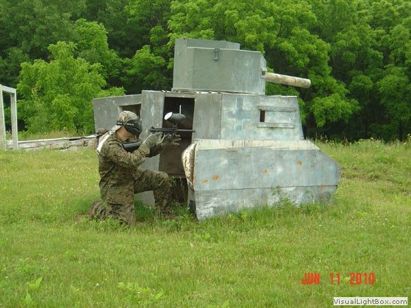 Soldier crouching behind a gray armored vehicle in a grassy field with trees nearby