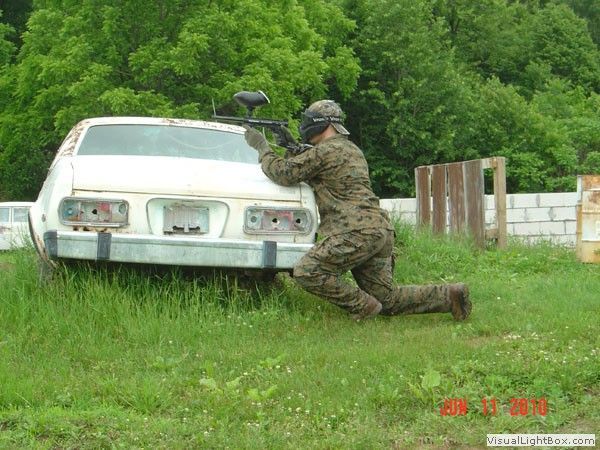 Soldier kneels behind a white car, aiming a firearm in a grassy field near trees.