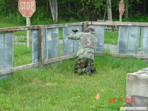 Person in camouflage kneels behind a paintball barrier at an outdoor field with stop signs.