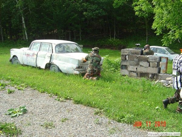 Man kneels by an old white car beside stacked cinder blocks in a grassy wooded area