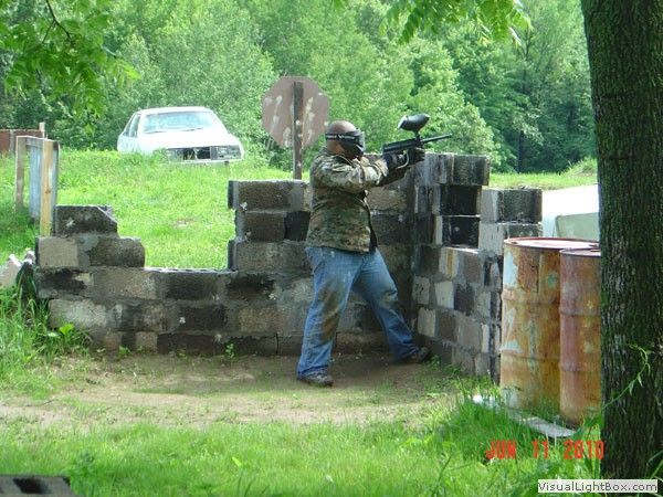Person in camouflage aiming a paintball marker from behind a brick barrier in a wooded outdoor field