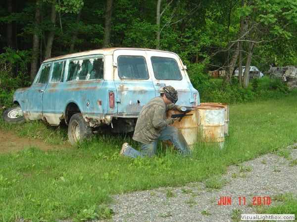 Person kneels by a rusty blue station wagon in a grassy yard with trees behind it