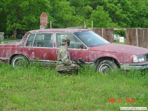 Soldier kneeling beside a maroon sedan in tall grass, with a stop sign and trees behind.