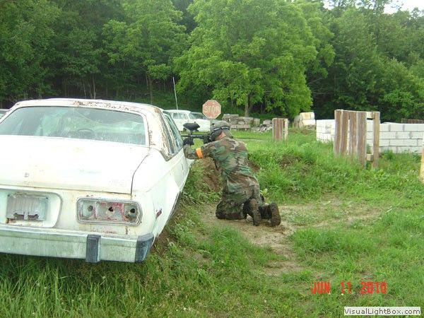 Camouflaged person crouches behind a white car, aiming a rifle in a grassy wooded area.
